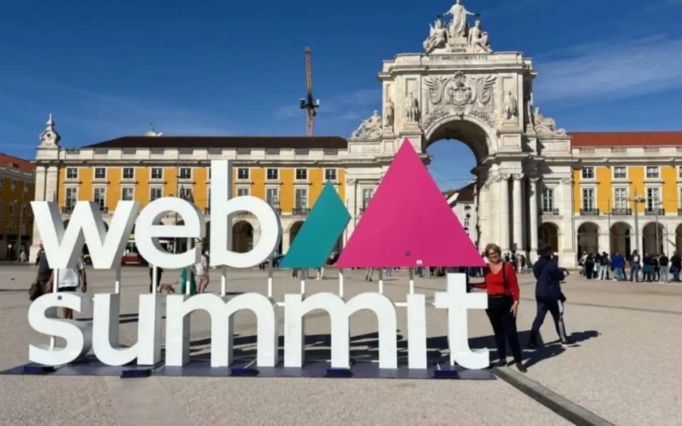 Large 3D Web Summit sign in front of the Arco da Rua Augusta in Praça do Comércio, Lisbon, with people around and a clear blue sky.